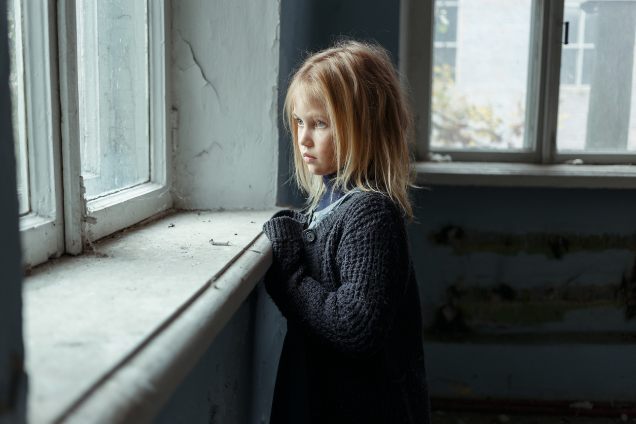 Hopeless life. Close up of depressed poor little girl standing near window